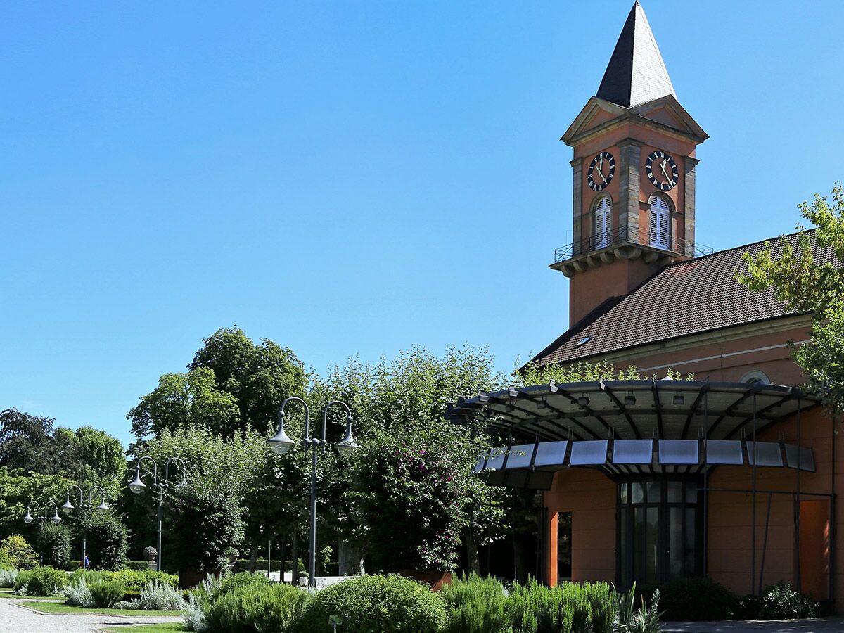Ein roter Glockenturm mit Uhr steht neben einer modernen Gebäudestruktur, umgeben von einer grünen Parkanlage unter blauem Himmel.