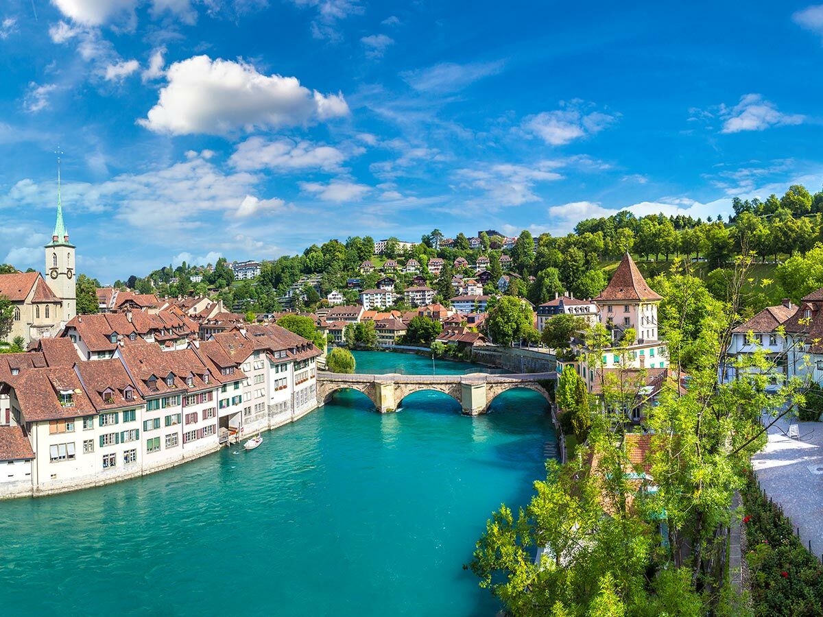Blick auf die Berner Altstadt mit historischen Gebäuden, dem türkisfarbenen Aare Fluss, Brücken und grünen Hügeln.