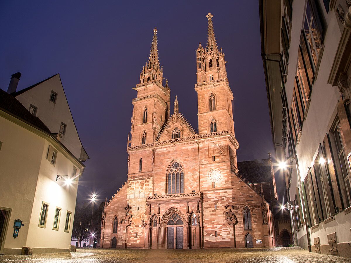 Imposantes Basler Münster bei Nacht, beleuchtet, mit zwei Türmen und historischen Gebäuden auf Kopfsteinpflasterplatz.