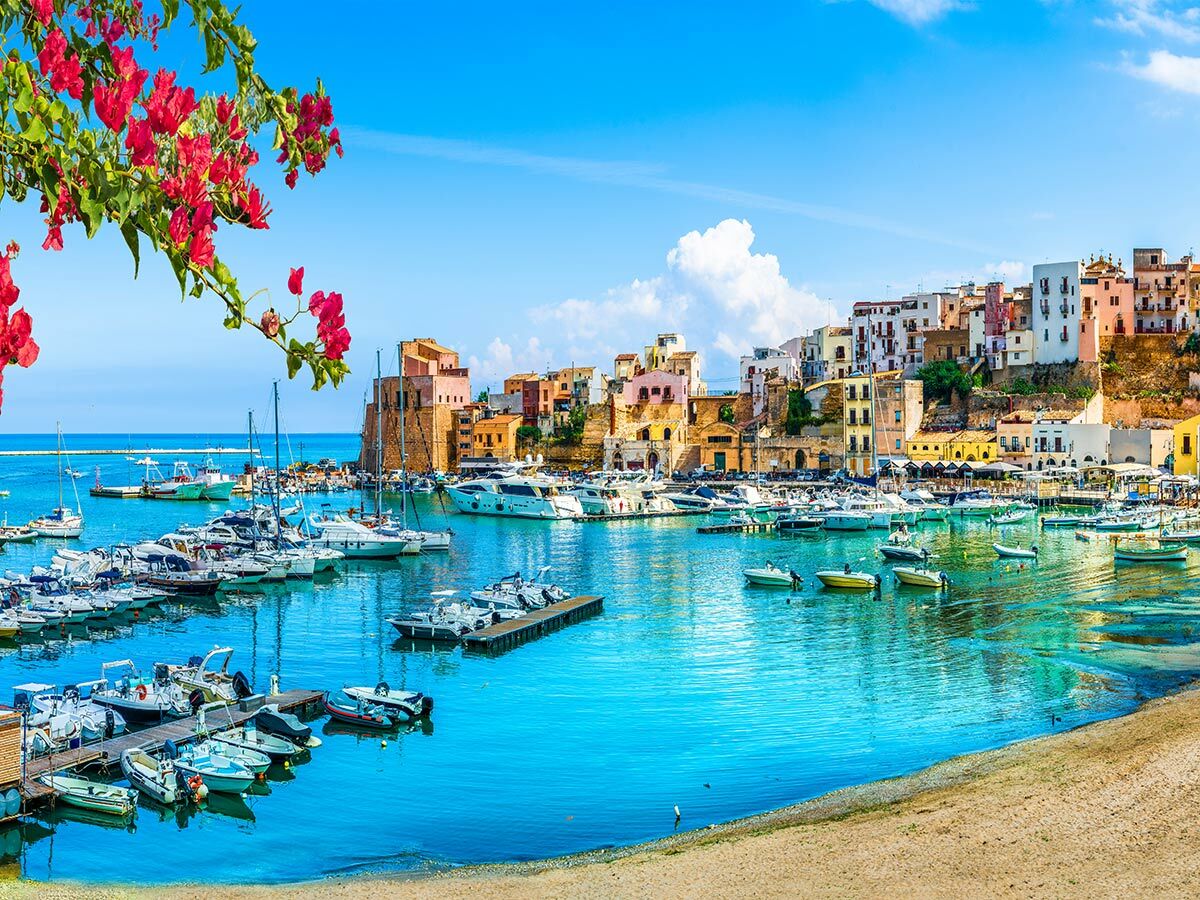 Malerischer italienischer Hafen mit vielen Booten, bunten Häusern an der Küste, klarem blauem Meer und Sandstrand unter strahlendem Himmel, umrahmt von roten Blüten.