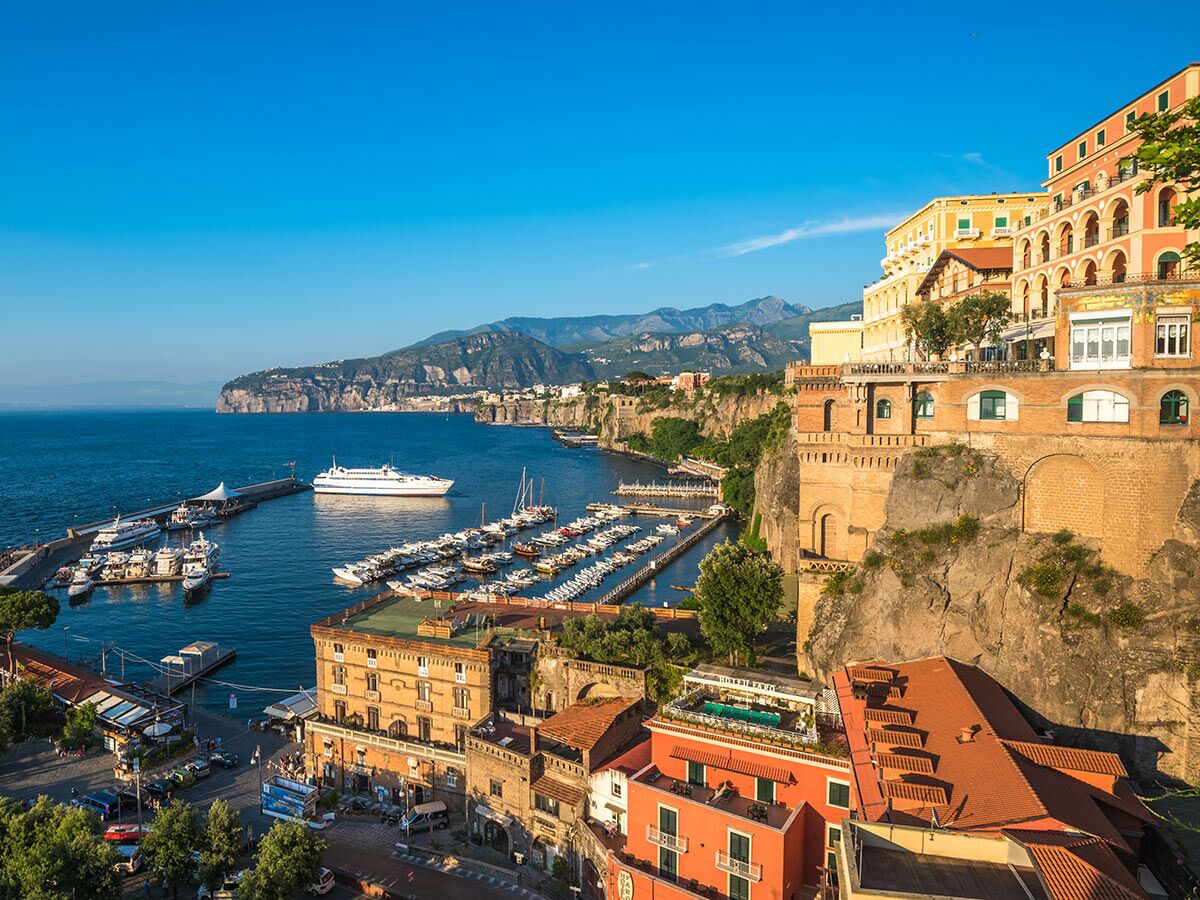 Malerische Küstenstadt Sorrento mit blauem Meer, belebtem Hafen voller Boote und farbenfrohen Gebäuden an steilen Klippen unter blauem Himmel.