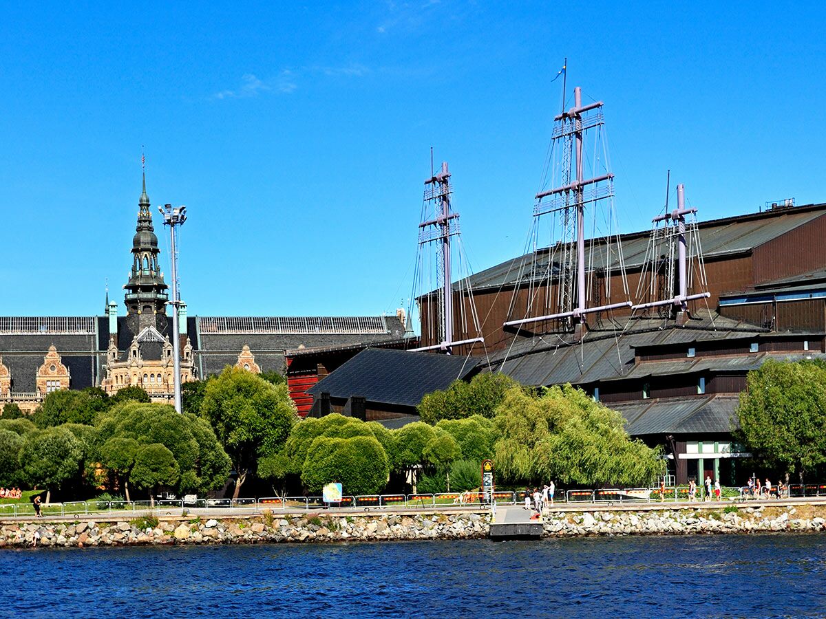 Blick auf das Vasa Museum mit seinen Masten und das Nordiska museet in Stockholm vom Wasser aus bei blauem Himmel.
