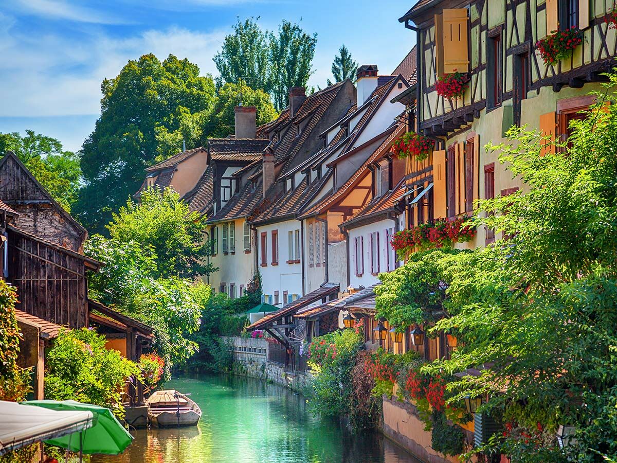 Idyllischer Kanal in Colmar mit bunten Fachwerkhäusern, blühenden Blumen und kleinen Booten unter blauem Himmel.