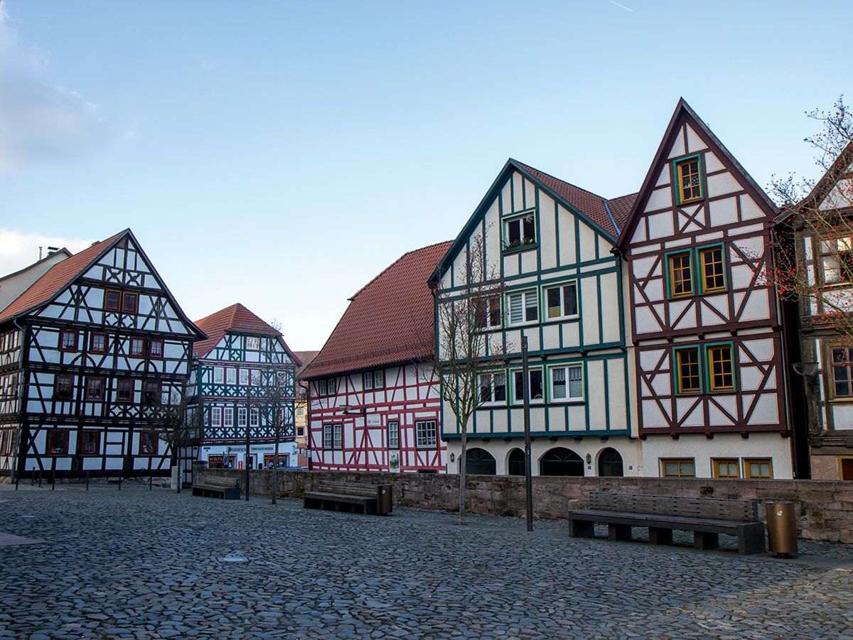 Mehrere bunte Fachwerkhäuser mit roten Dächern und Holzbalken stehen an einem leeren Kopfsteinpflaster-Marktplatz in einer deutschen Altstadt.