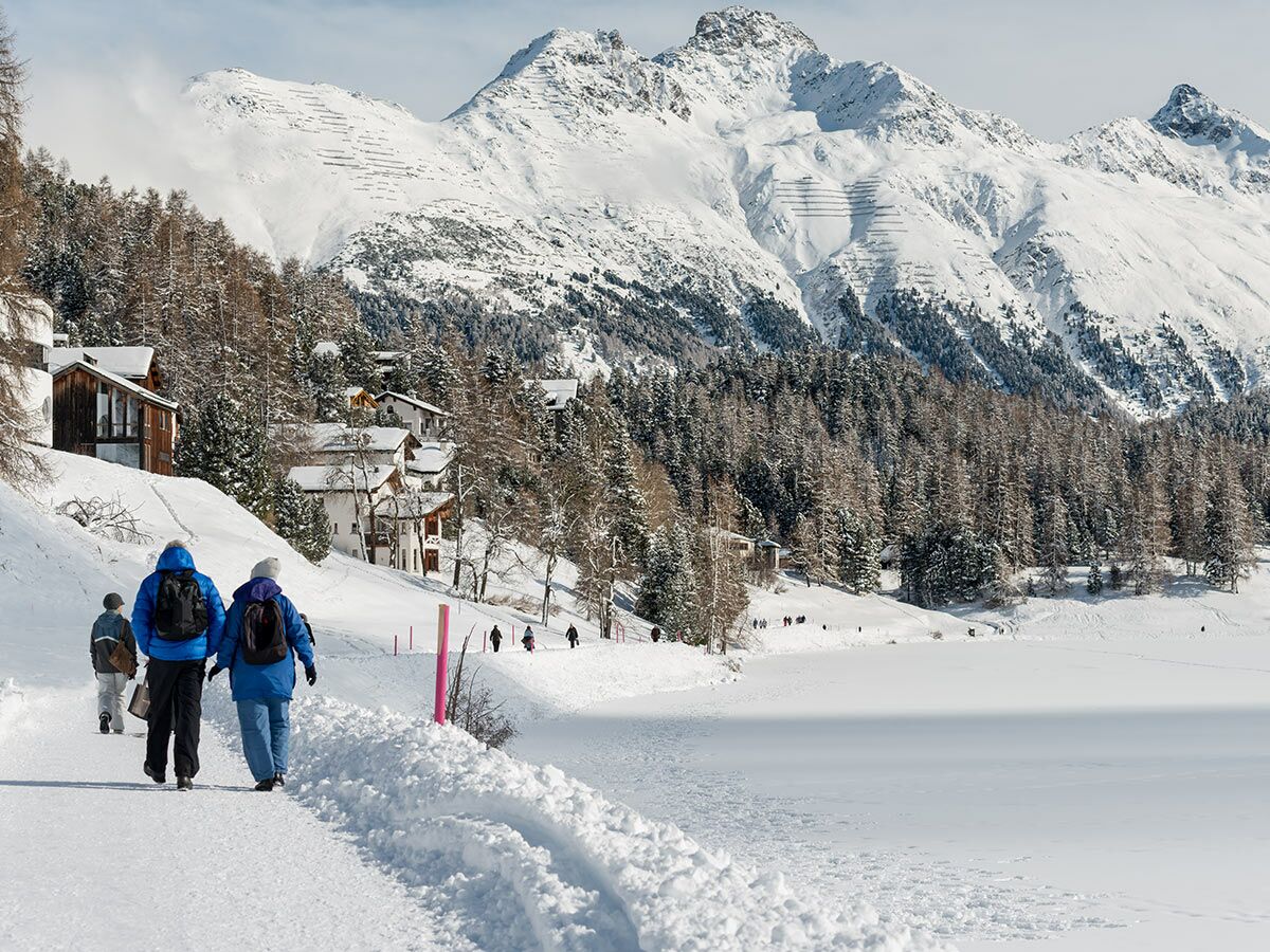Zwei Wanderer gehen auf einem verschneiten Weg am Ufer eines zugefrorenen Sees, mit Alpendorf und Bergen im Hintergrund.
