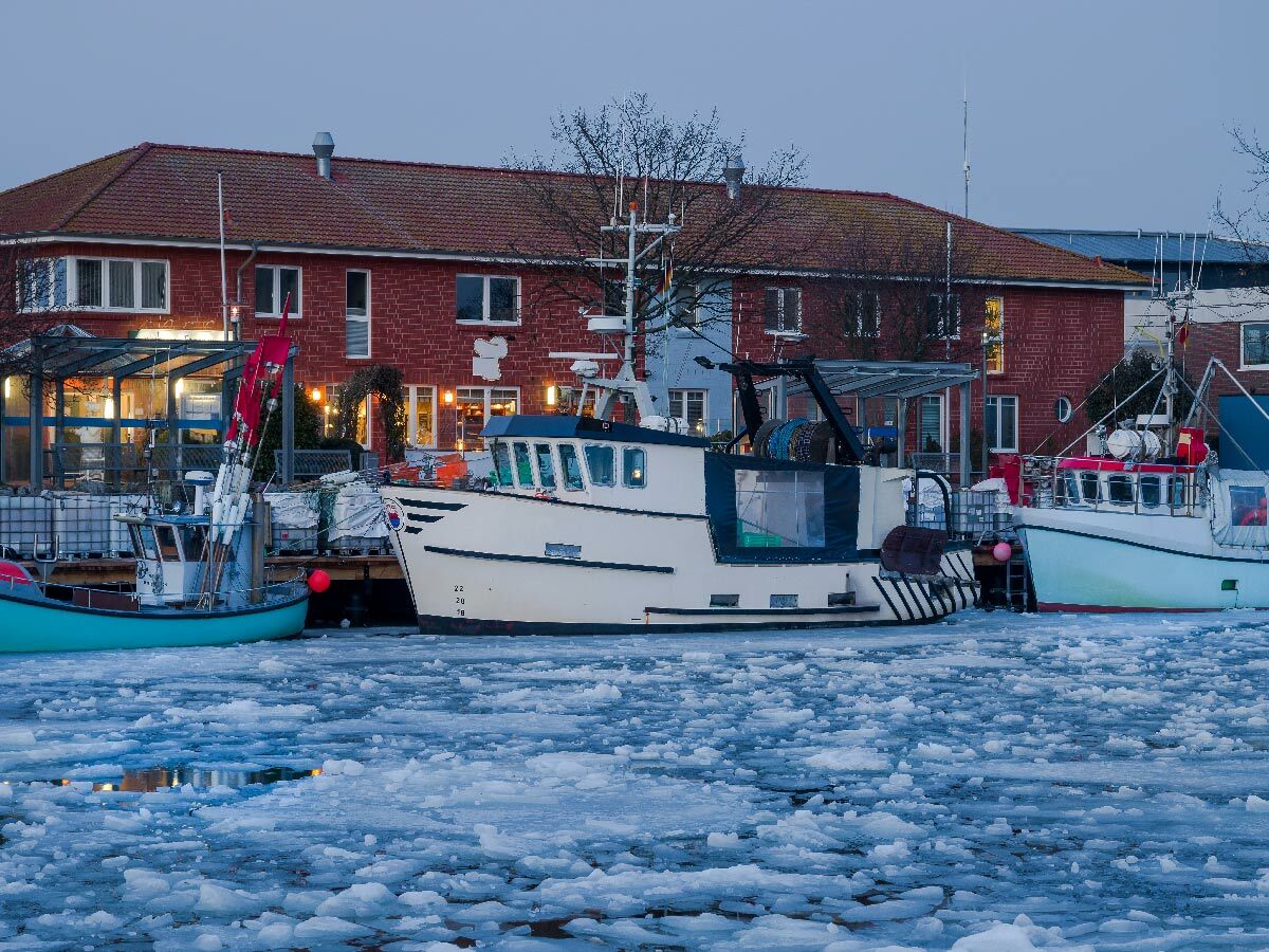 Mehrere Fischerboote liegen im winterlichen Hafen bei Dämmerung. Das Wasser ist mit Eisschollen bedeckt, im Hintergrund ein Backsteinhaus.