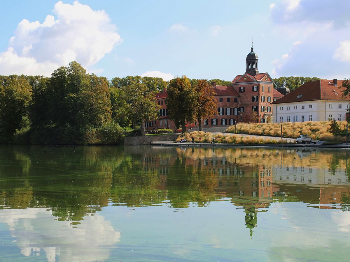 Malerischer Blick auf ein historisches Backsteinschloss mit Turm, im ruhigen See spiegelnd, umgeben von Herbstbäumen unter blauem Himmel.