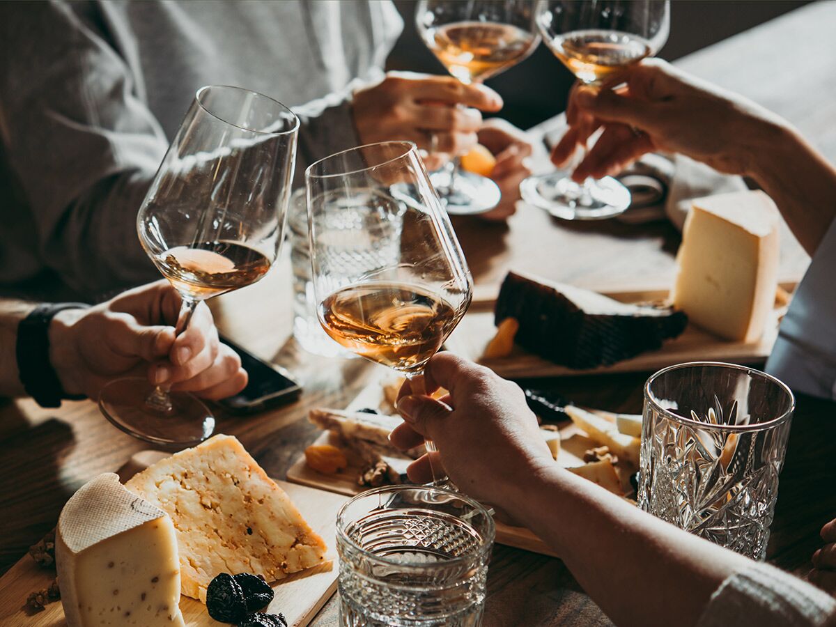 Mehrere Personen halten Weingläser bei einer Wein- und Käseverkostung mit verschiedenen Käsesorten, Trockenfrüchten und Nüssen auf Holztisch.
