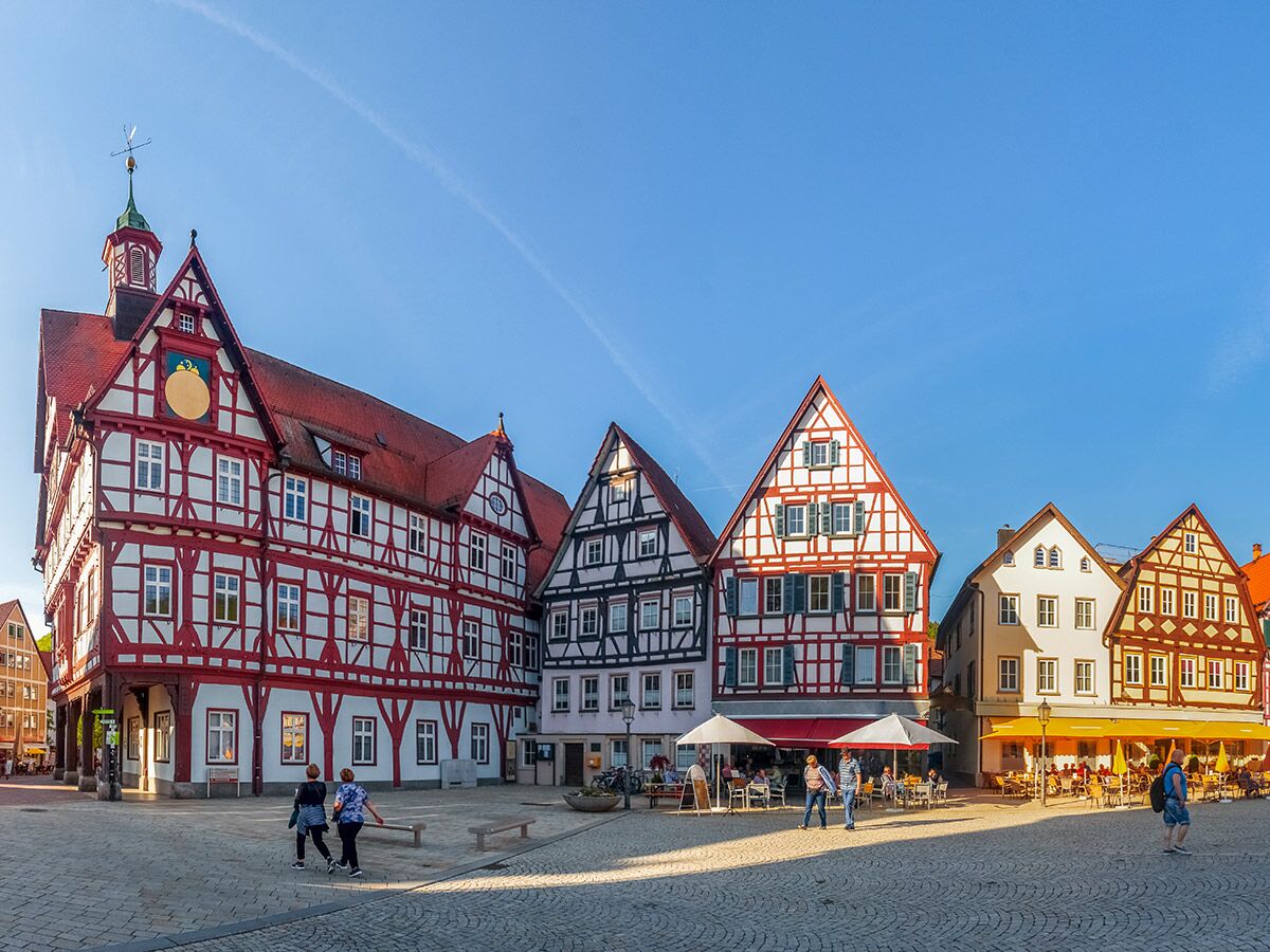 Sonniger Marktplatz mit roten Fachwerkhäusern, Rathaus und Cafés in einer deutschen Altstadt, Menschen genießen den Tag.
