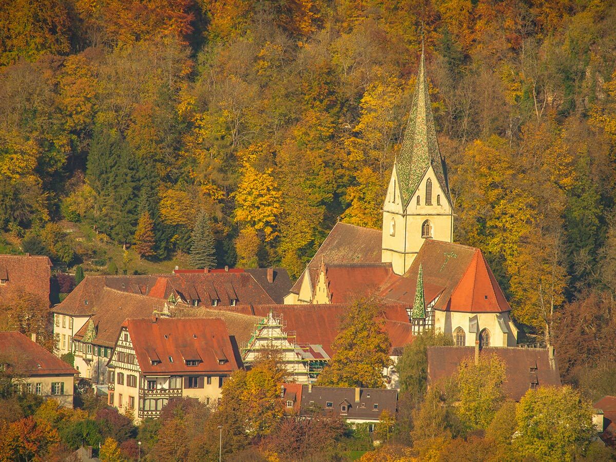 Malerisches Kloster Blaubeuren im goldenen Herbst mit historischen Gebäuden, rotem Dach und hohem Kirchturm, umgeben von bunten Bäumen.