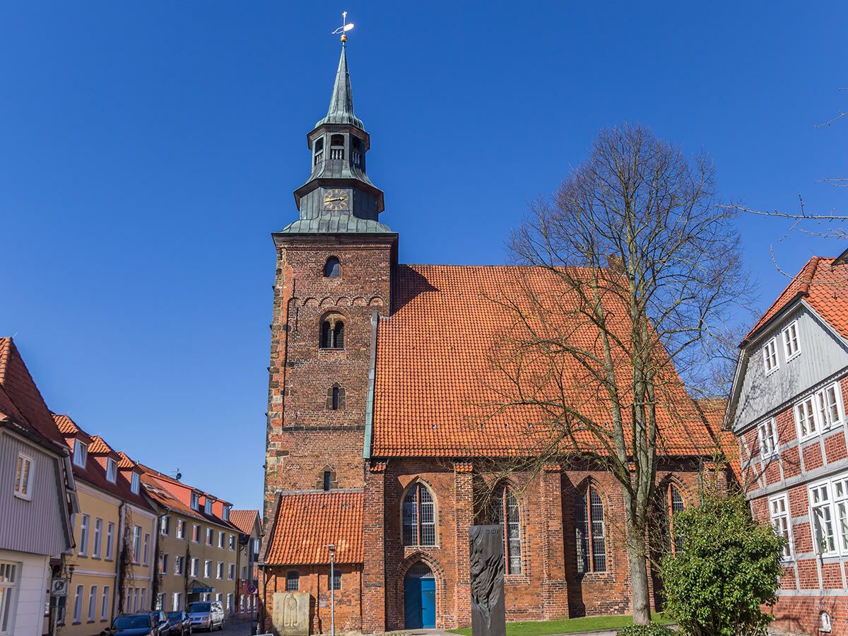 Blick auf eine historische Backsteinkirche mit hohem Turm und Uhr unter blauem Himmel, umgeben von alten Häusern in einer europäischen Altstadt.
