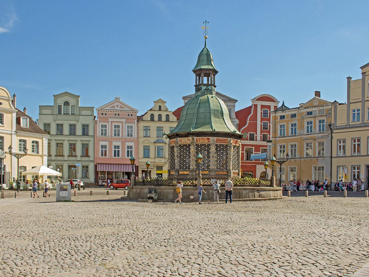 Historischer Marktplatz Wismar mit Wasserkunst Brunnen, umgeben von bunten Giebelhäusern unter blauem Himmel.
