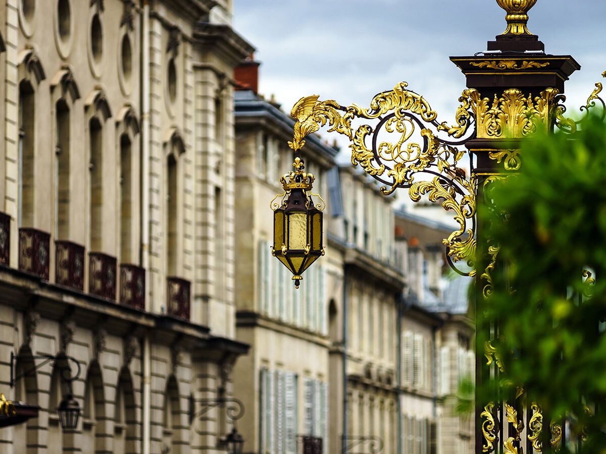 Detail einer vergoldeten, verzierten Straßenlaterne vor einer historischen europäischen Häuserzeile mit Balkonen und Fenstern.
