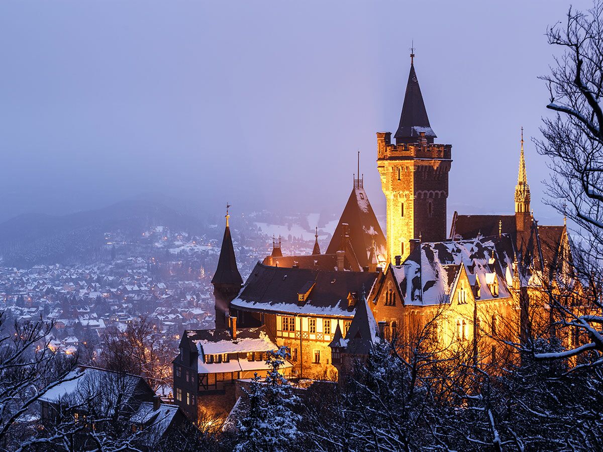 Schloss Wernigerode im Winter bei Dämmerung, hell beleuchtet mit Schnee auf den Dächern, dahinter verschneite Stadt.