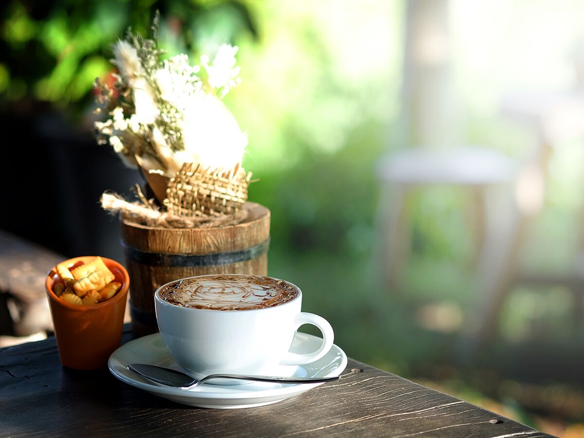 Eine Tasse Kaffee mit Latte Art steht auf einem rustikalen Holztisch neben Crackern und einer Blumen-Deko im Freien.