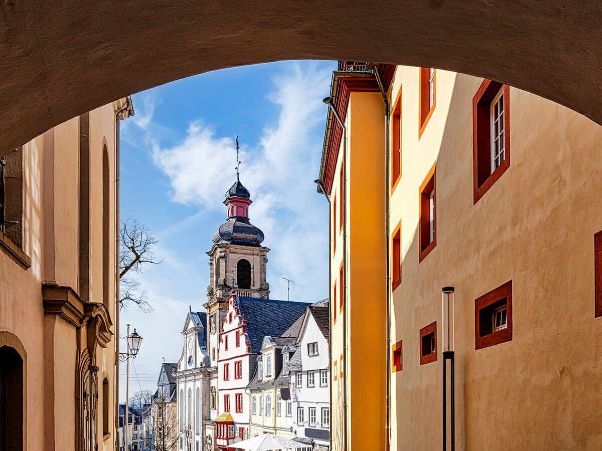 Blick durch einen steinernen Bogen auf eine historische Gasse mit barocker Kirche, farbigen Häusern und blauem Himmel in einer deutschen Altstadt.