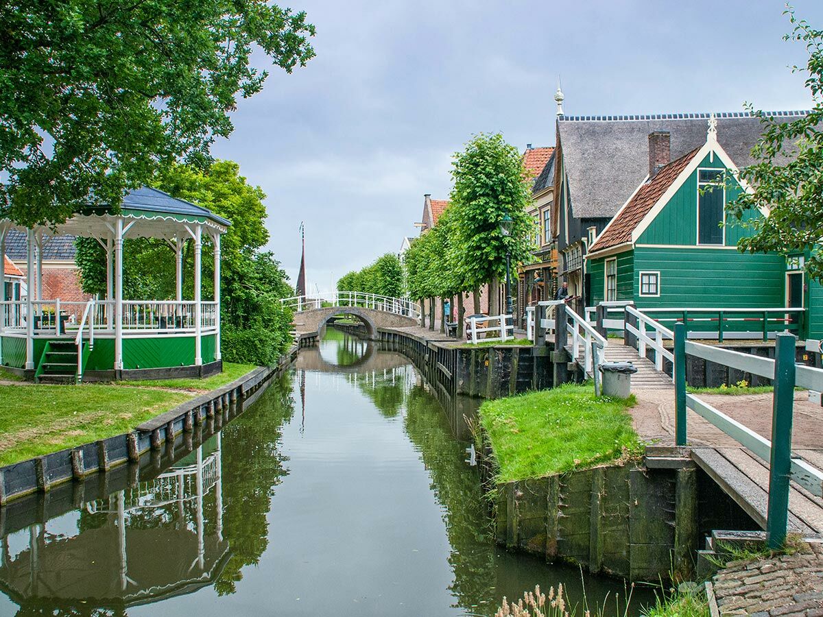 Blick auf einen Kanal in einem holländischen Dorf mit grünen Holzhäusern, einem Pavillon und einer Steinbrücke unter bewölktem Himmel.