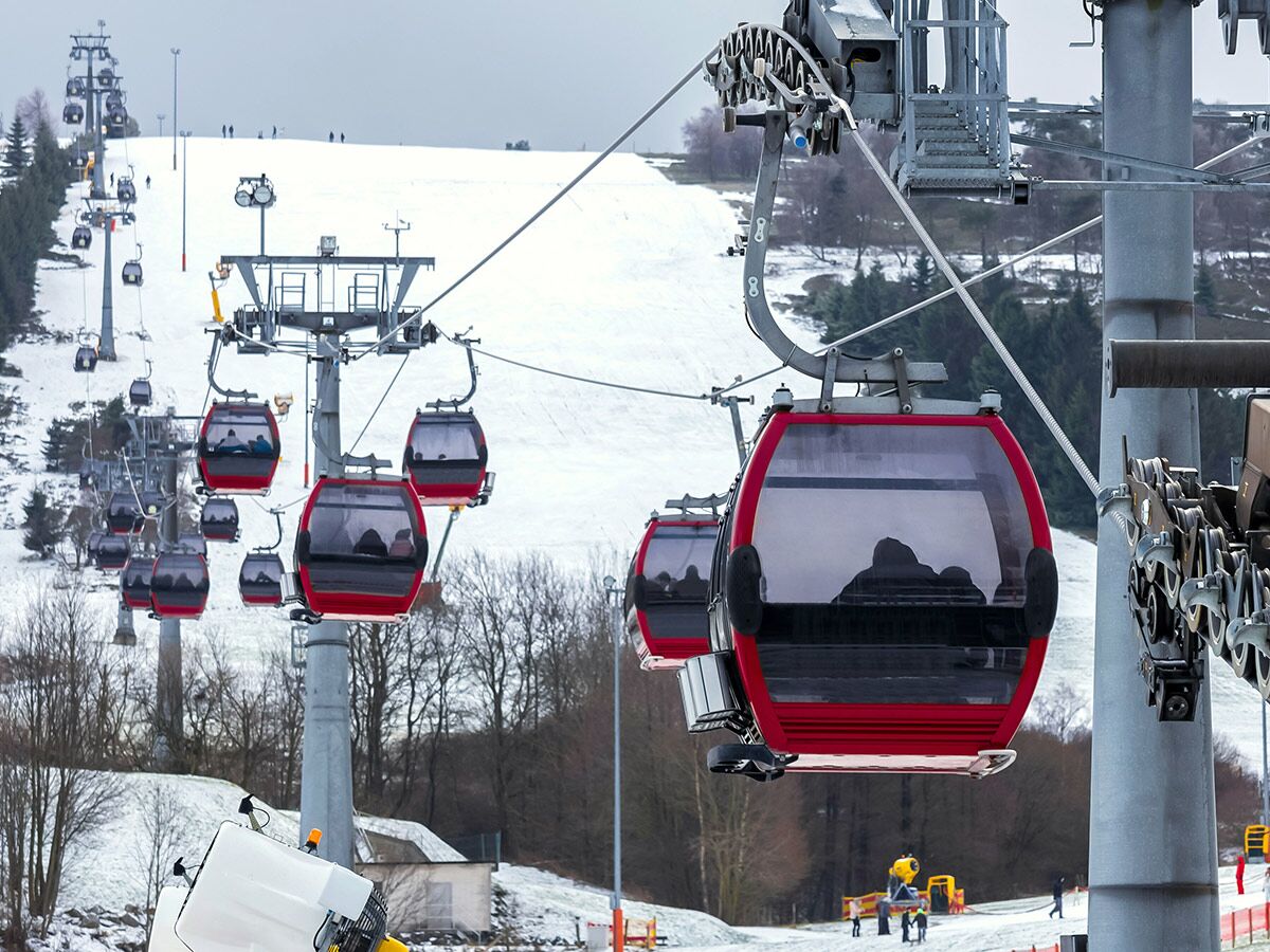 Rote Gondelbahnkabinen transportieren Skifahrer einen verschneiten Berg in einem belebten Winterskigebiet unter bewölktem Himmel hinauf.