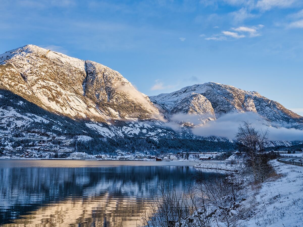 Panorama einer winterlichen Fjordlandschaft mit schneebedeckten Bergen, ruhigem Wasser und einem Dorf am Ufer.