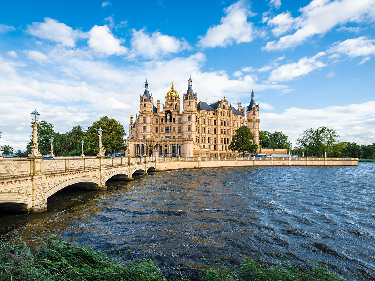 Majestätisches Schweriner Schloss mit goldener Kuppel und Türmen am See, verbunden durch eine elegante Steinbrücke unter blauem Himmel.