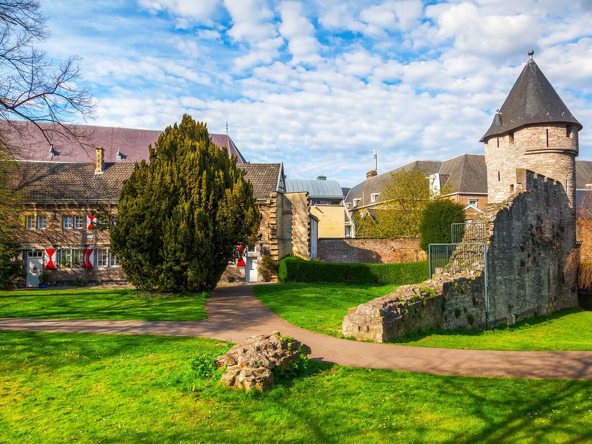 Sonniger Park mit mittelalterlichem Burgturm, alten Steinmauern, einem großen Baum und historischen Gebäuden mit rot-weißen Fensterläden.