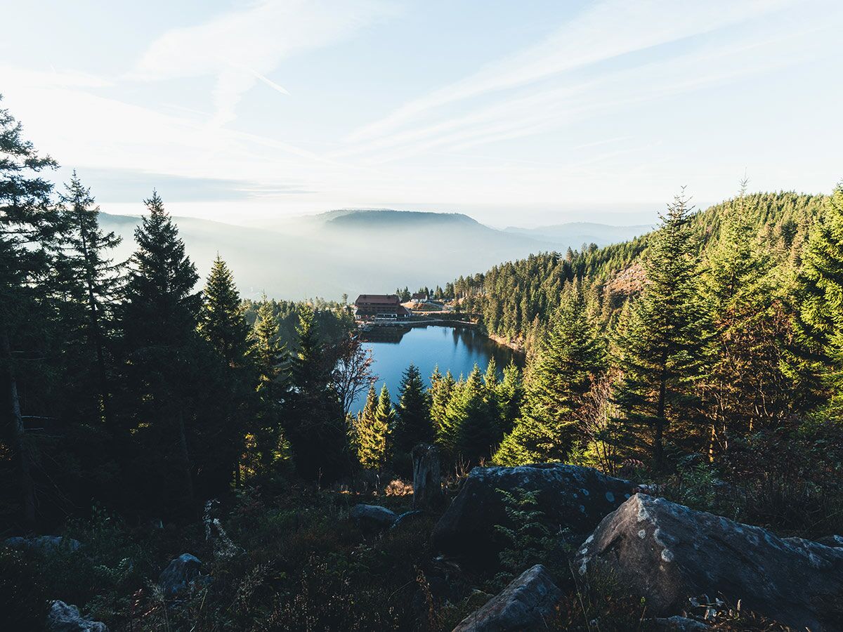 Malerischer Bergsee mit Hütte am Ufer, umgeben von dichten Nadelwäldern und nebligen Bergen im Hintergrund.