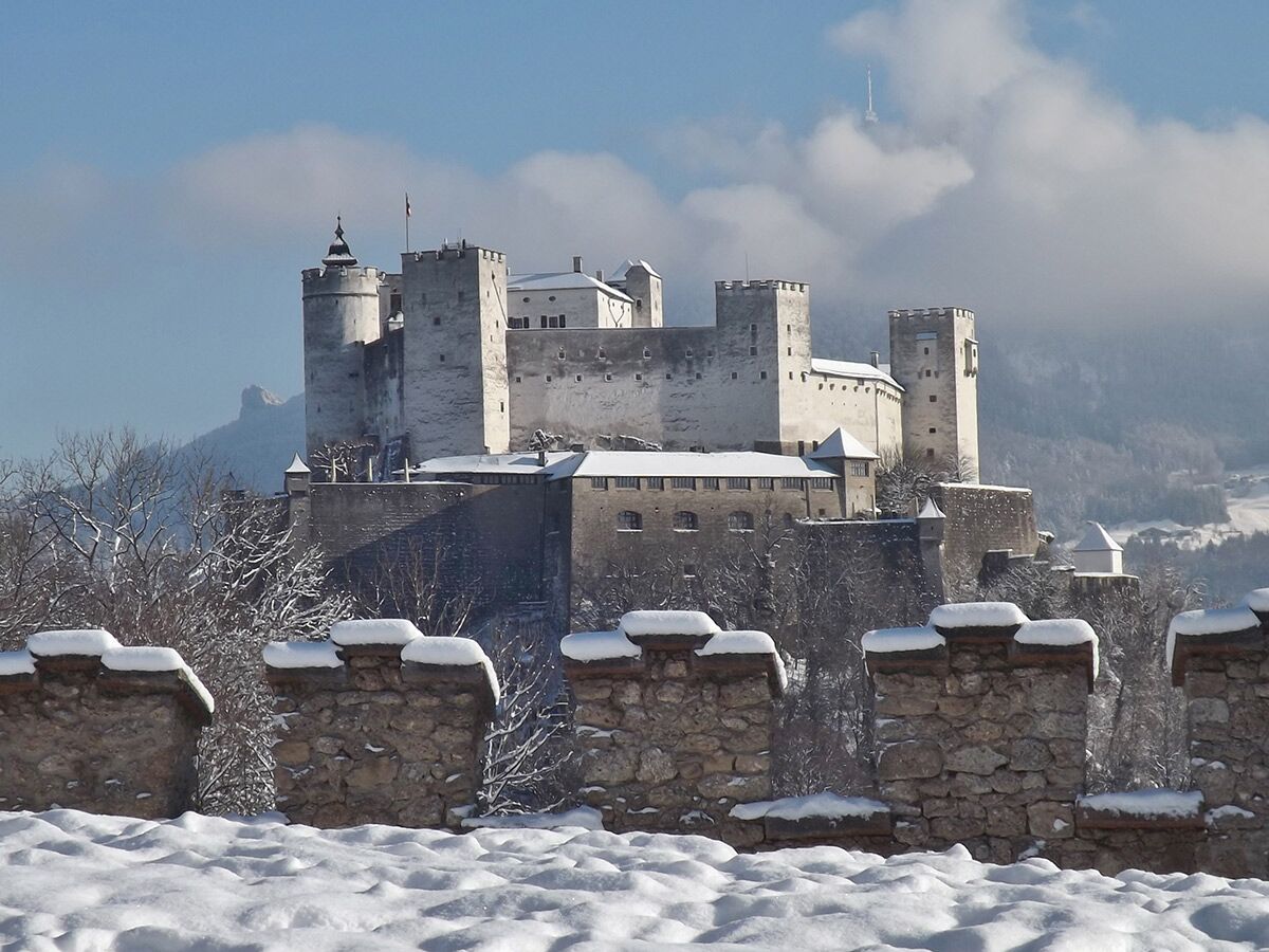 Die Hohensalzburg Festung in Salzburg, Österreich, im Winter mit schneebedeckten Dächern, Mauern und Bergen unter blauem Himmel.