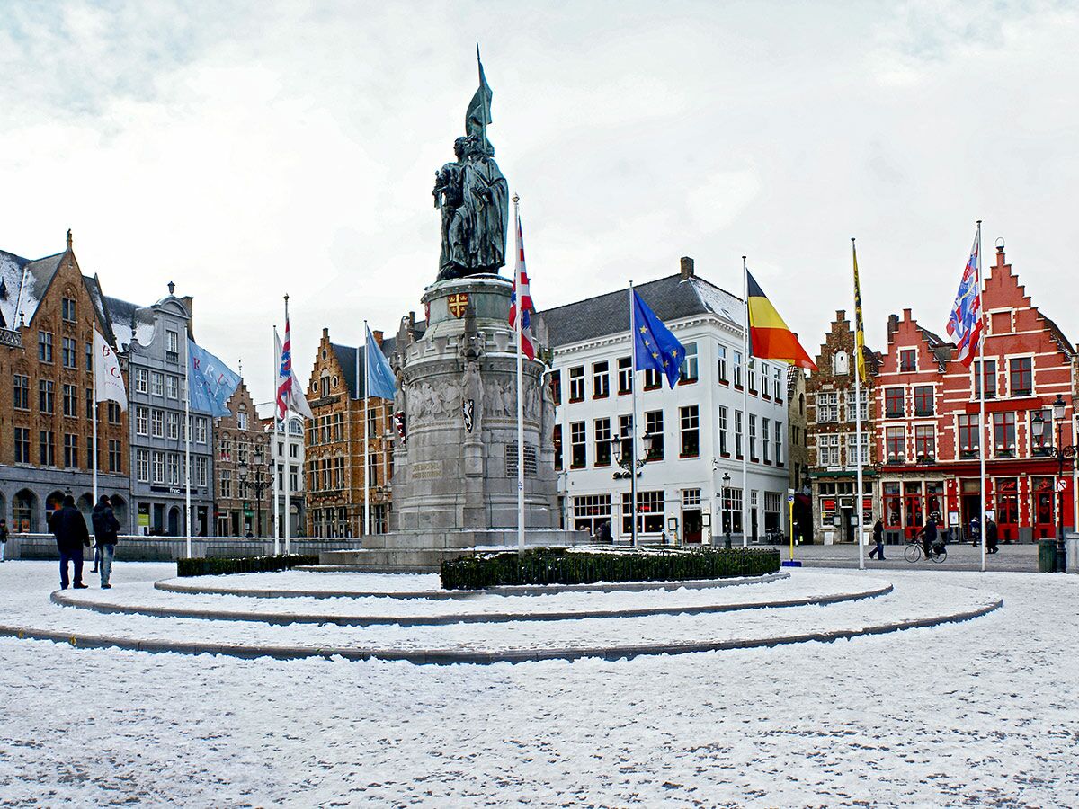 Verschneiter Grote Markt Brügge mit Breydel und de Coninck Denkmal, umgeben von historischen Giebelhäusern und wehenden Fahnen.