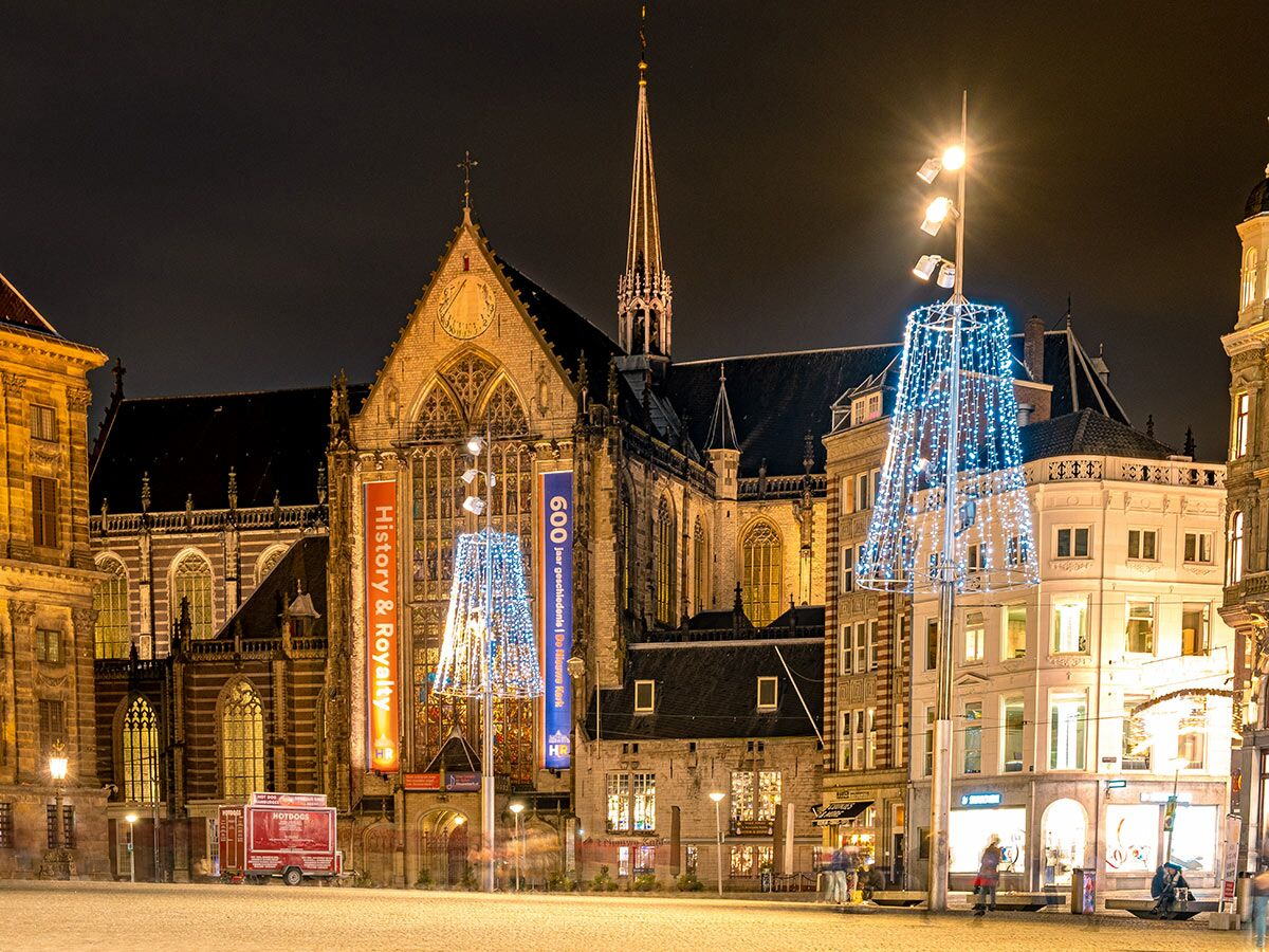 Belebter Dam Platz Amsterdam bei Nacht mit festlich beleuchteter Nieuwe Kerk, Weihnachtsdeko, Gebäuden und Foodtruck.