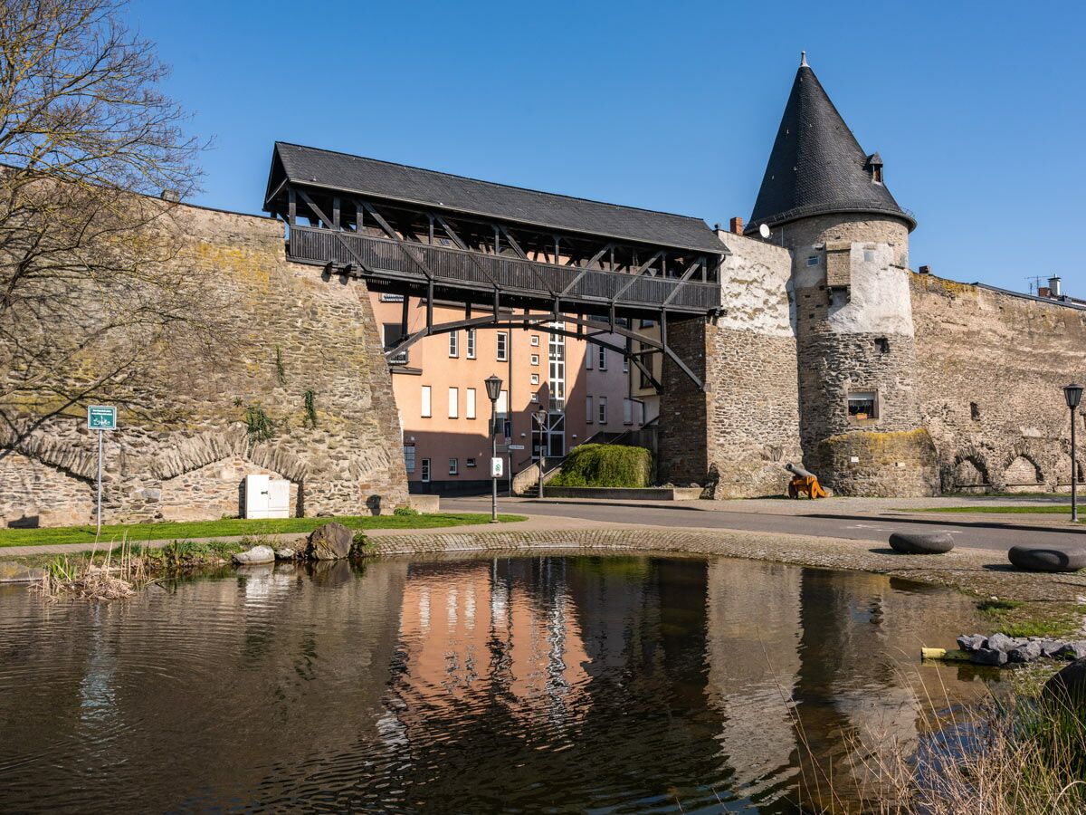 Historische Stadtmauer mit überdachter Holzbrücke, rundem Turm, Kanone, Teich und blauer Himmel an einem sonnigen Tag.