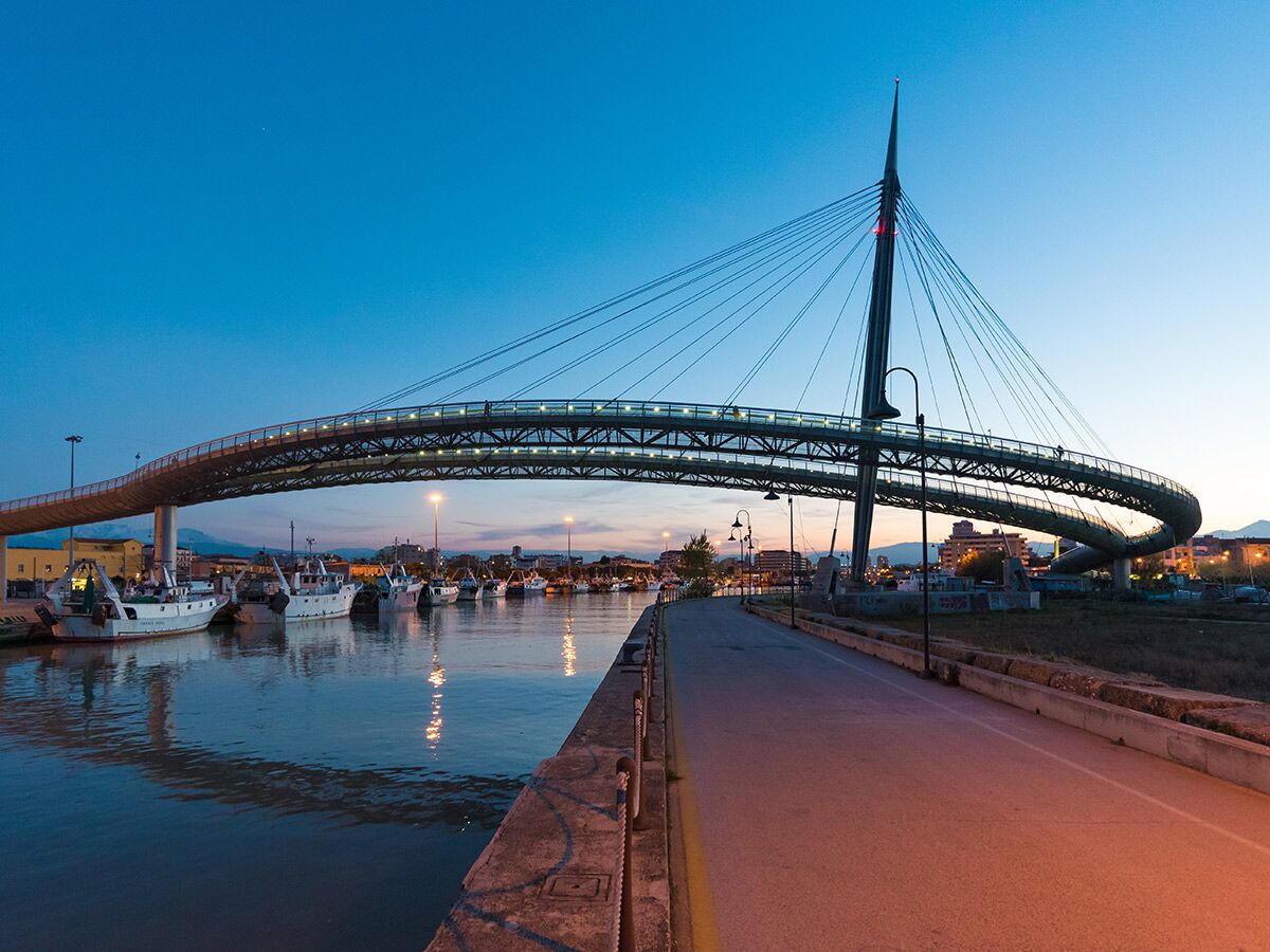 Moderne geschwungene Fußgängerbrücke Ponte del Mare in Pescara bei Dämmerung mit Fischerbooten im Fluss.