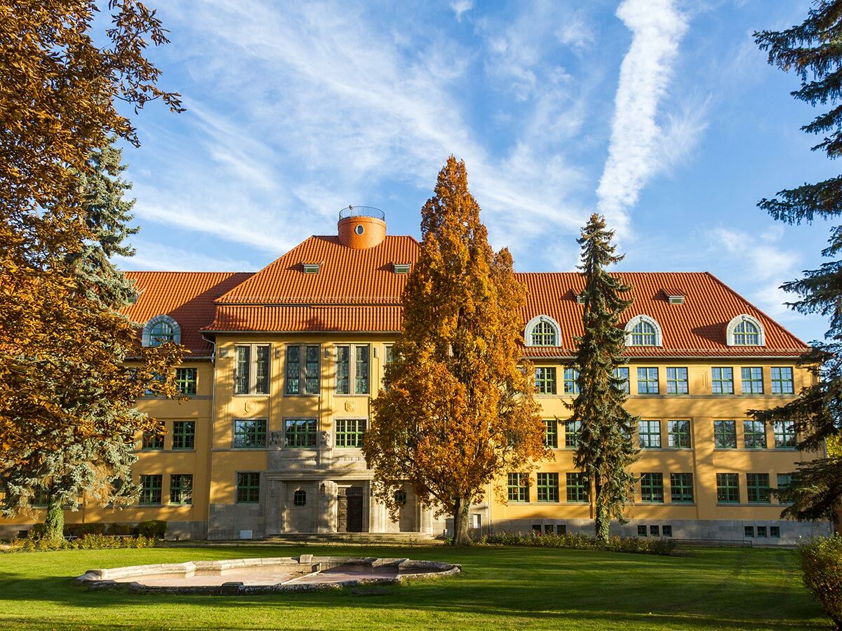 Großes, gelbes Gebäude mit rotem Ziegeldach, umgeben von Herbstbäumen und grünem Rasen mit Teich unter blauem Himmel.