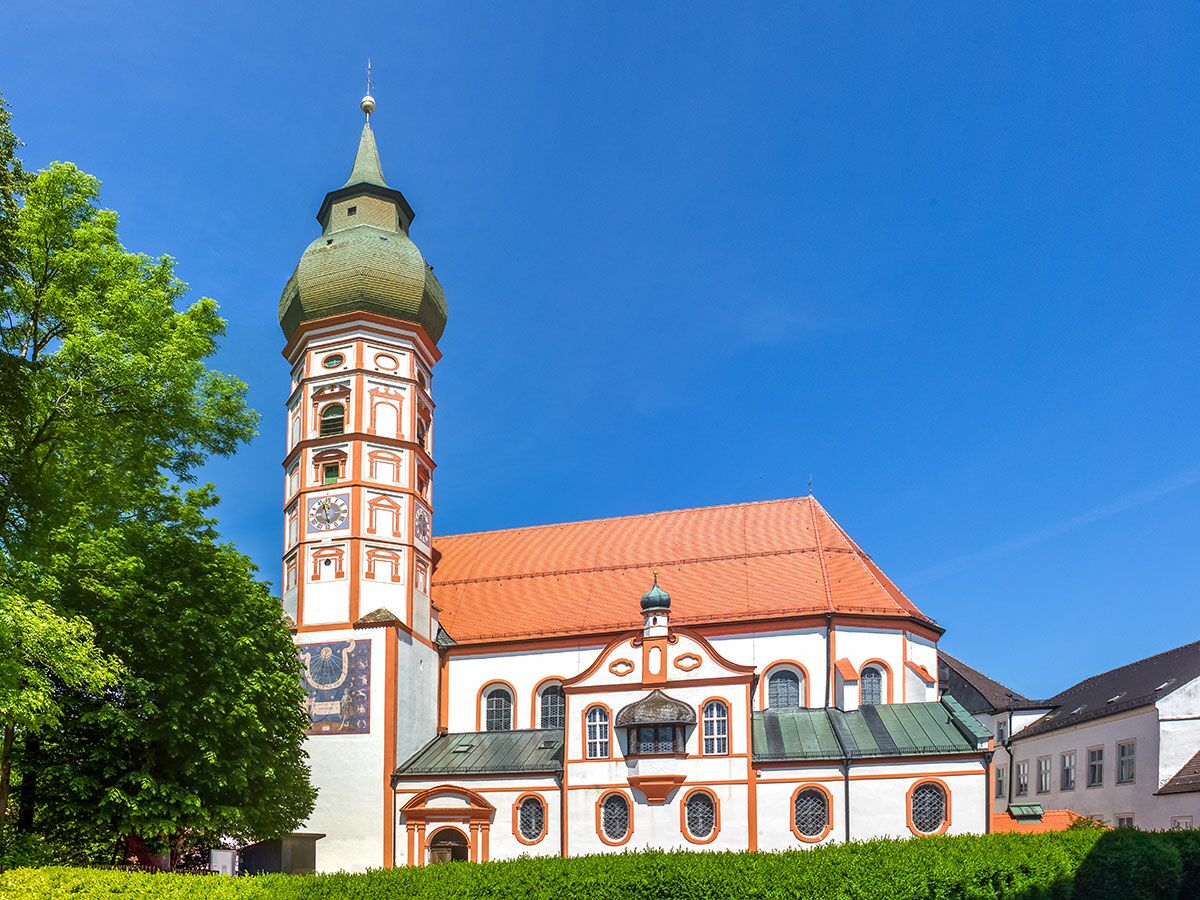 Barocke Abtei mit hohem Turm, Zwiebelkuppel, roten Dächern und einer Sonnenuhr unter blauem Himmel.
