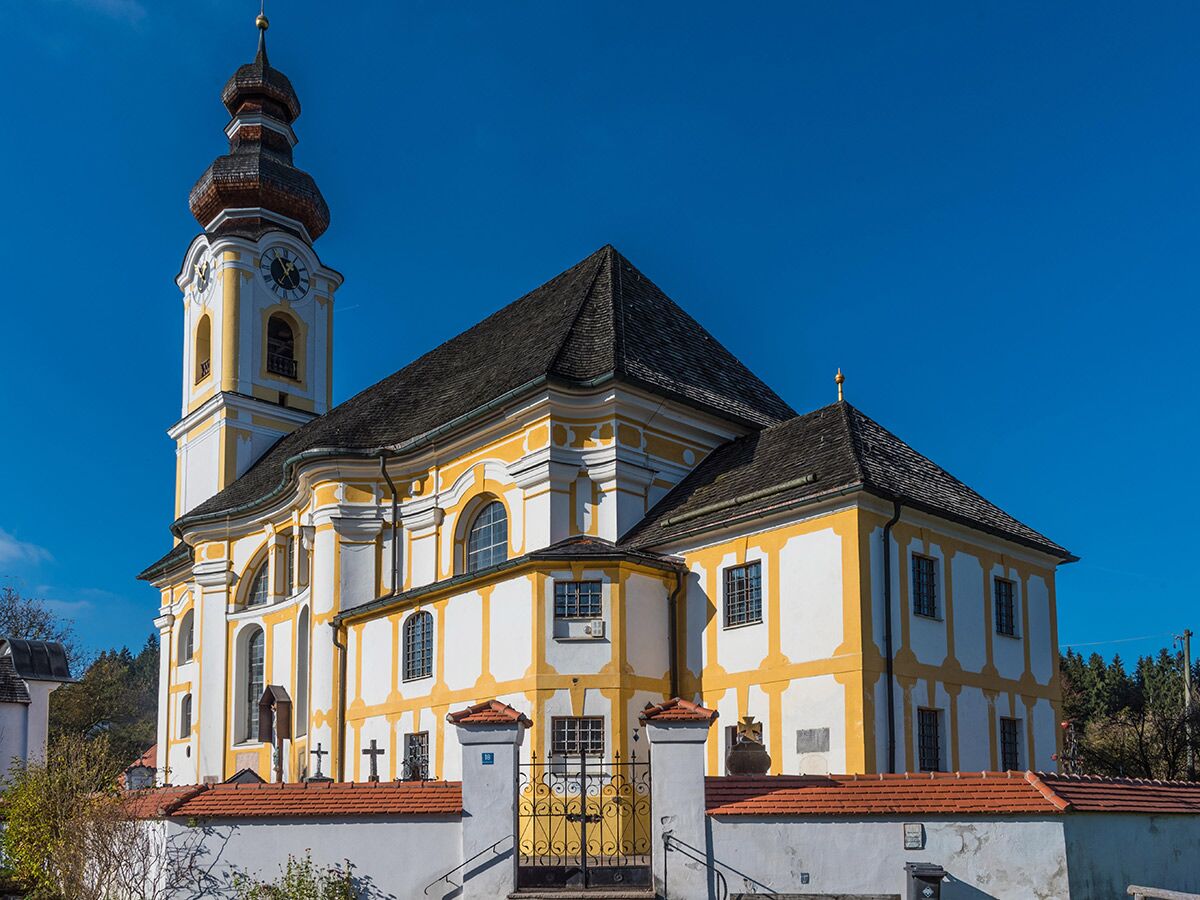 Prächtige barocke Kirche mit gelb-weißer Fassade, hohem Glockenturm mit Zwiebelkuppel und Uhren vor blauem Himmel.