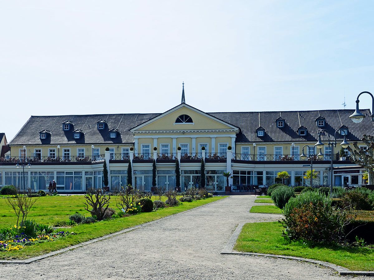 Elegantes Hotelgebäude mit gelber Fassade, dunklem Dach und Dachgauben. Grüner Garten mit Blumenbeeten und Kiesweg im Frühling.