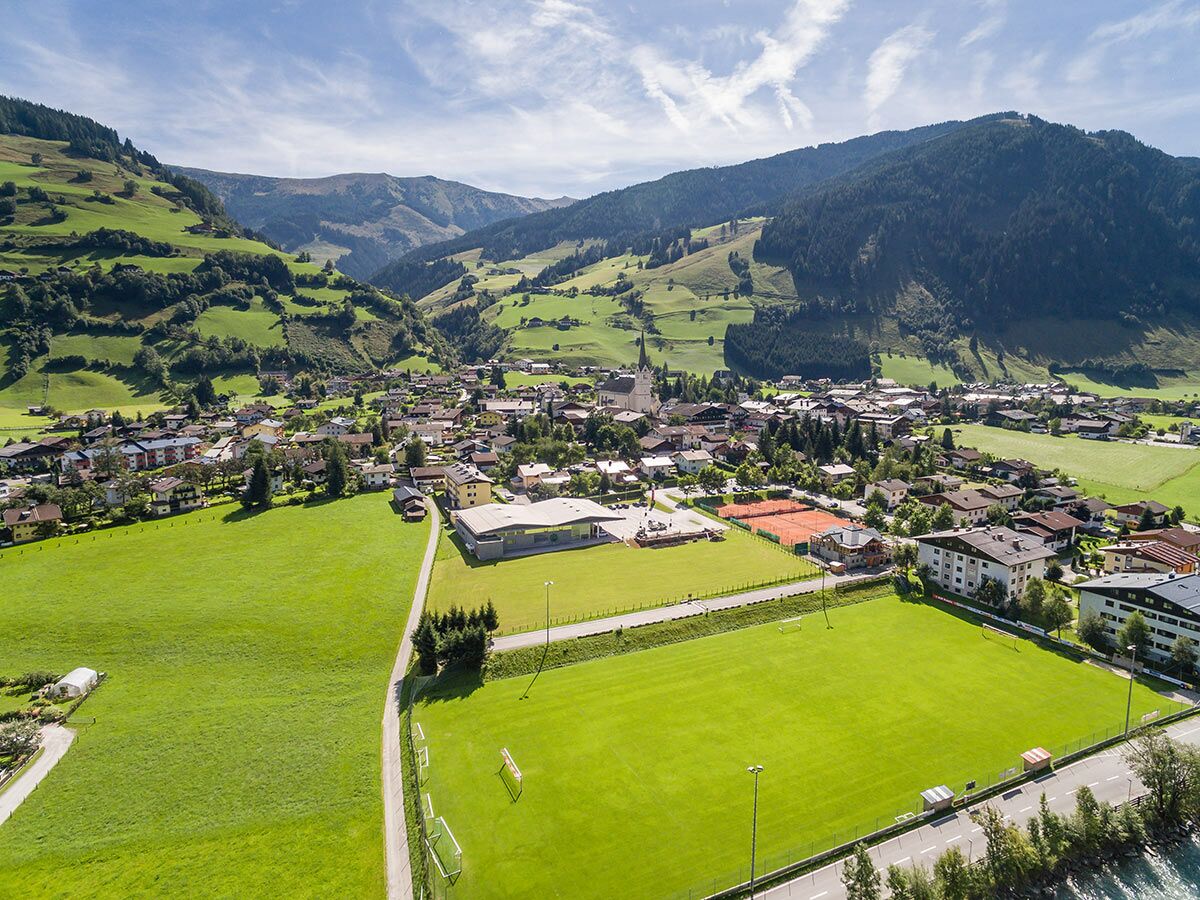 Luftaufnahme eines idyllischen Bergdorfes in den Alpen mit Kirche, Häusern, grünen Wiesen, Sportplätzen und Bergen.