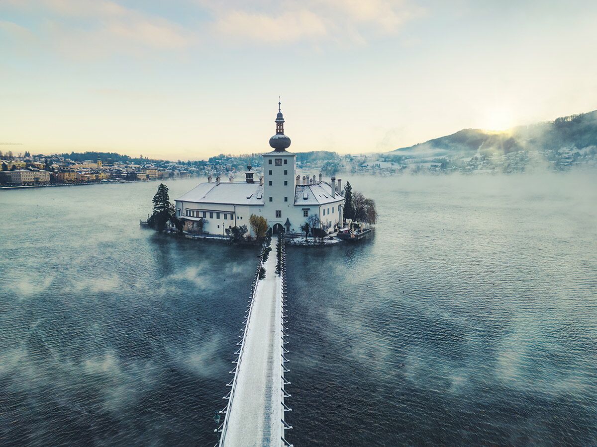 Schloss Ort Gmunden im nebligen Traunsee, mit verschneiter Brücke zur Insel, winterliche Landschaft und Stadt im Hintergrund.