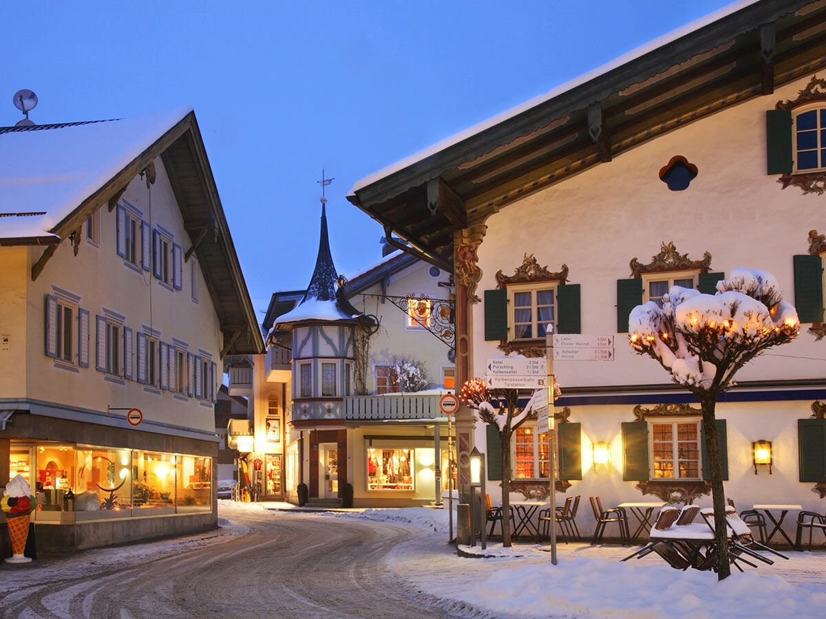 Ein winterlicher Abend in einer verschneiten bayerischen Alpenstadt mit traditionellen Häusern und beleuchteten Geschäften.