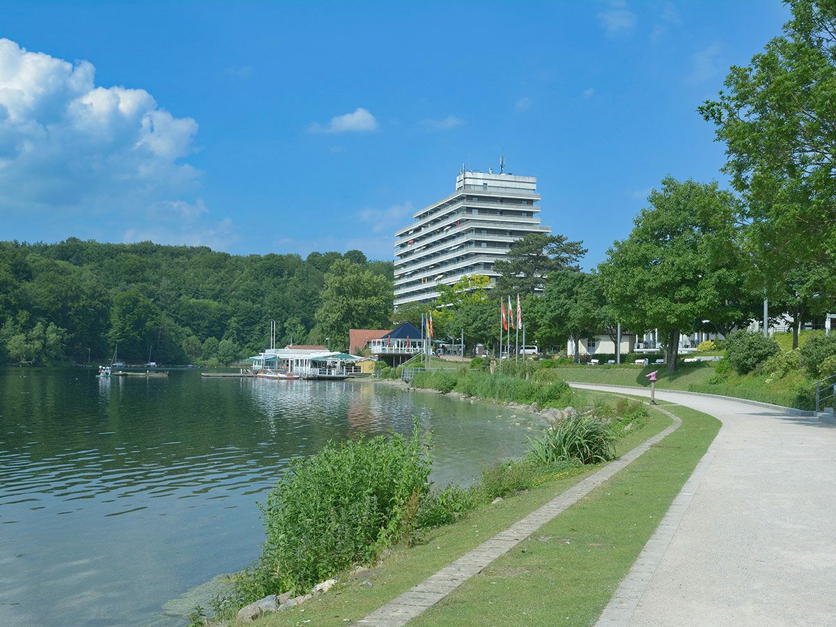 Blick auf ein ruhiges Seeufer mit einem modernen Gebäude, Restaurants und einer Promenade im Grünen.