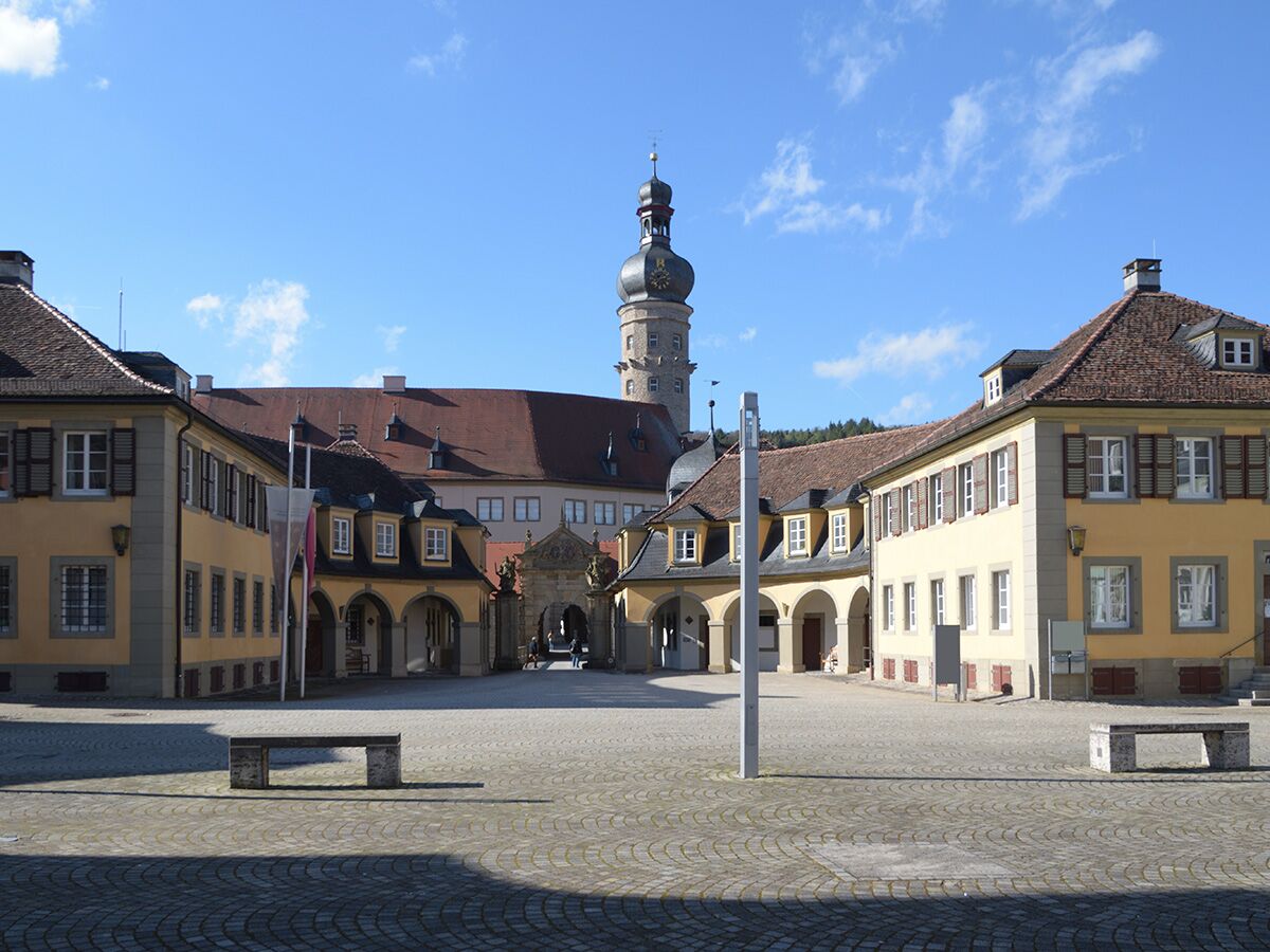 Historischer Schlosshof mit gelben Gebäuden, Kopfsteinpflaster, Steinbänken und einem hohen Uhrturm unter blauem Himmel.