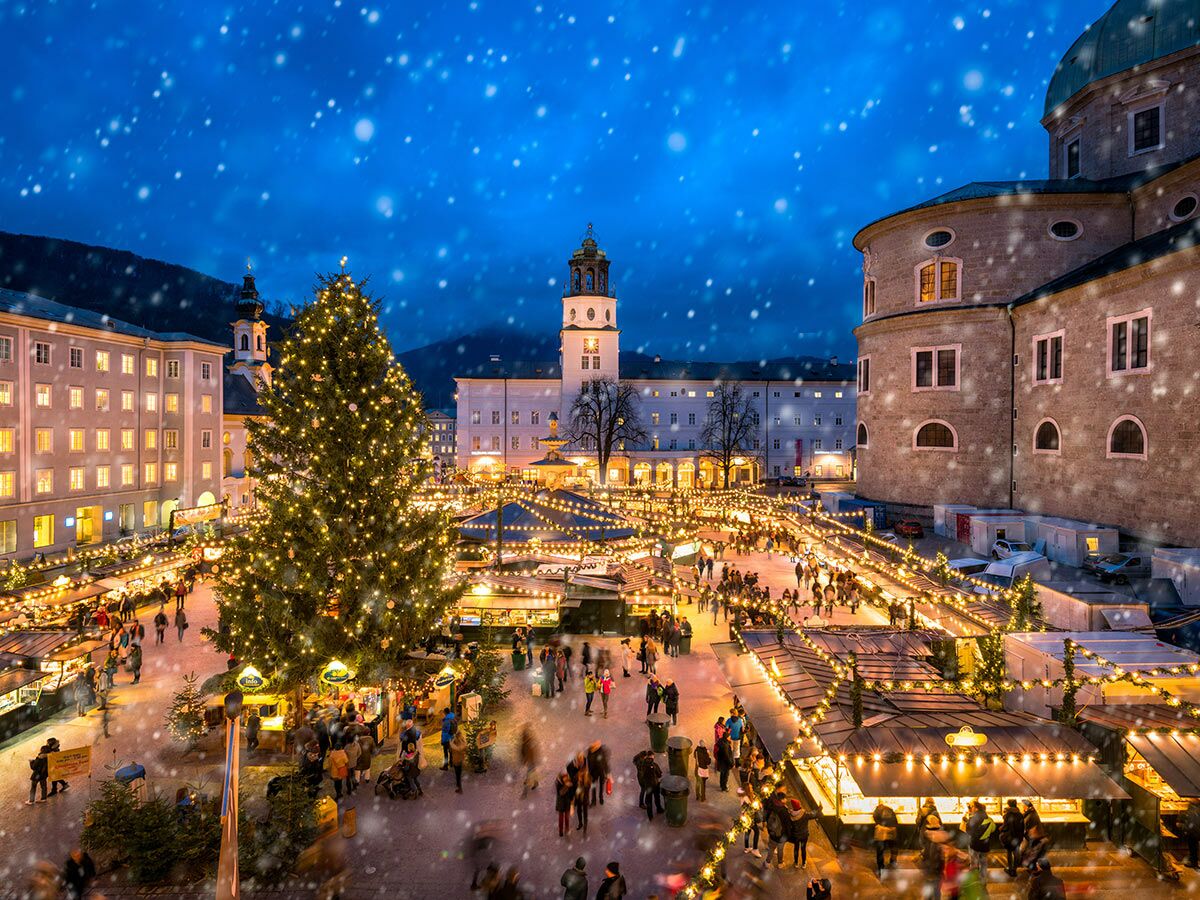 Blick von oben auf den festlichen Weihnachtsmarkt in Salzburg bei Schnee am Abend, mit beleuchtetem Christbaum und Marktständen.