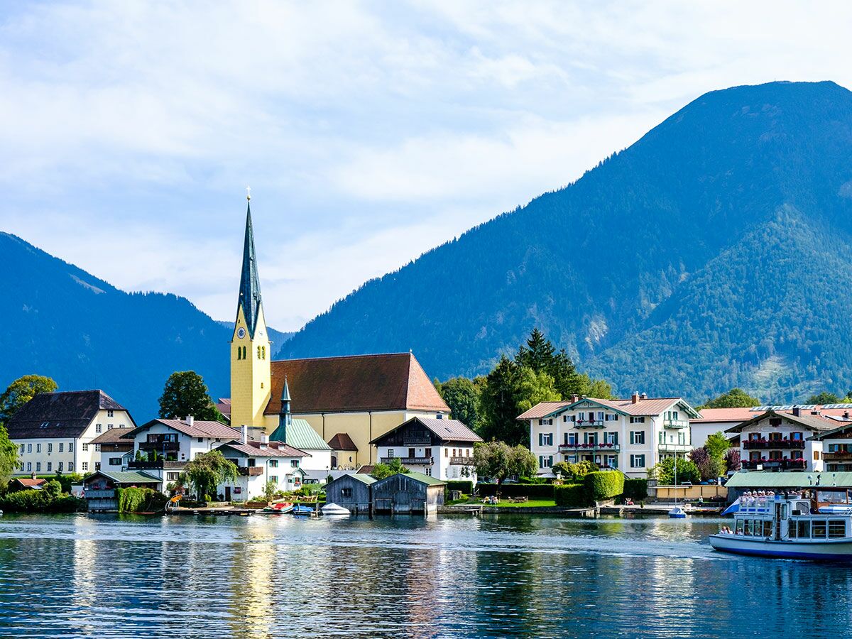 Malerisches bayerisches Dorf am Tegernsee mit Kirchturm, Häusern und Bergen. Boote auf blauem Wasser unter klarem Himmel.