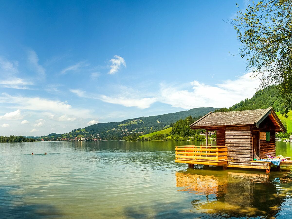 Malerische Holzhütte mit Steg auf einem klaren Bergsee. Grüne Berge, blauer Himmel, Wolken. Zwei Schwimmer im Wasser.