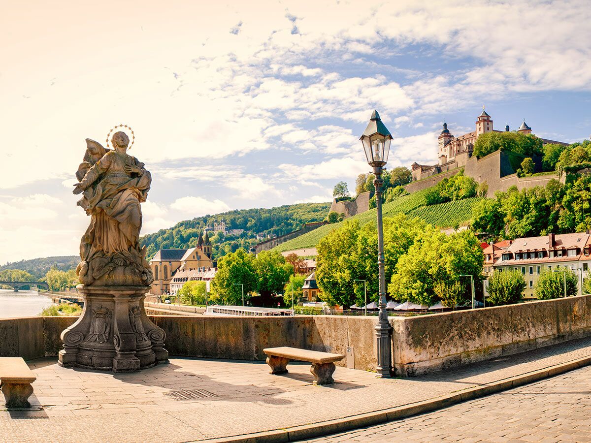 Historische Alte Mainbrücke Würzburg mit Statue, Fluss Main, Weinberg und Festung Marienberg unter blauem Himmel bei Sonnenschein.