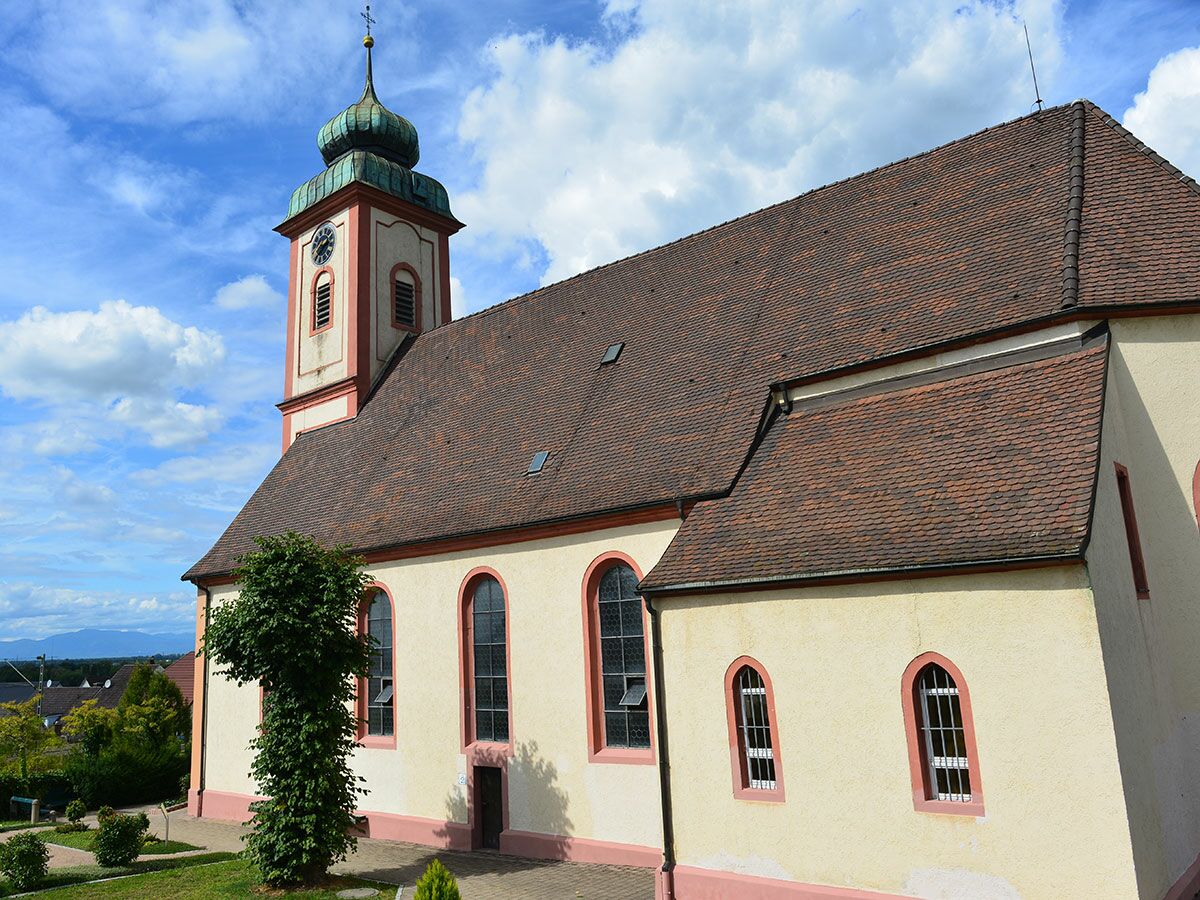 Seitenansicht einer historischen Kirche mit Zwiebelturm, Uhr, rotem Ziegeldach und Bogenfenstern unter blauem Himmel mit Wolken.