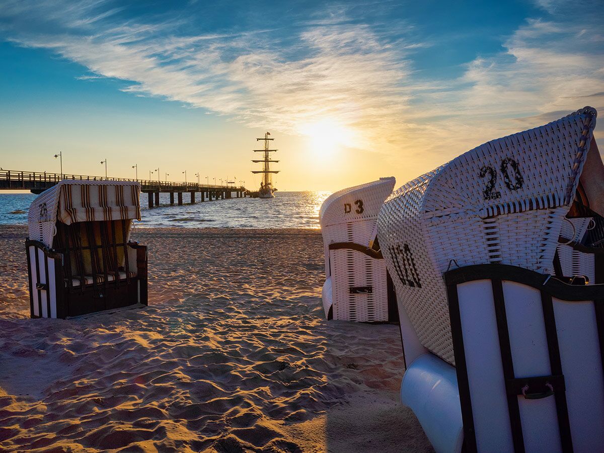 Sonnenuntergang am Strand mit Strandkörben, Seebrücke und einem Segelschiff im goldenen Licht des Himmels.