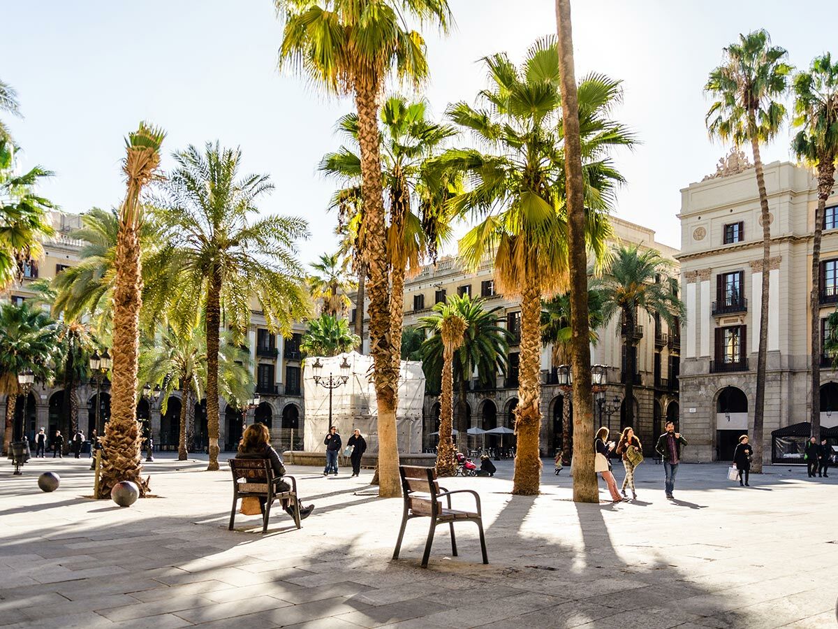 Sonniger Stadtplatz in Barcelona mit hohen Palmen, historischen Gebäuden, Menschen spazieren und sitzen auf Bänken.