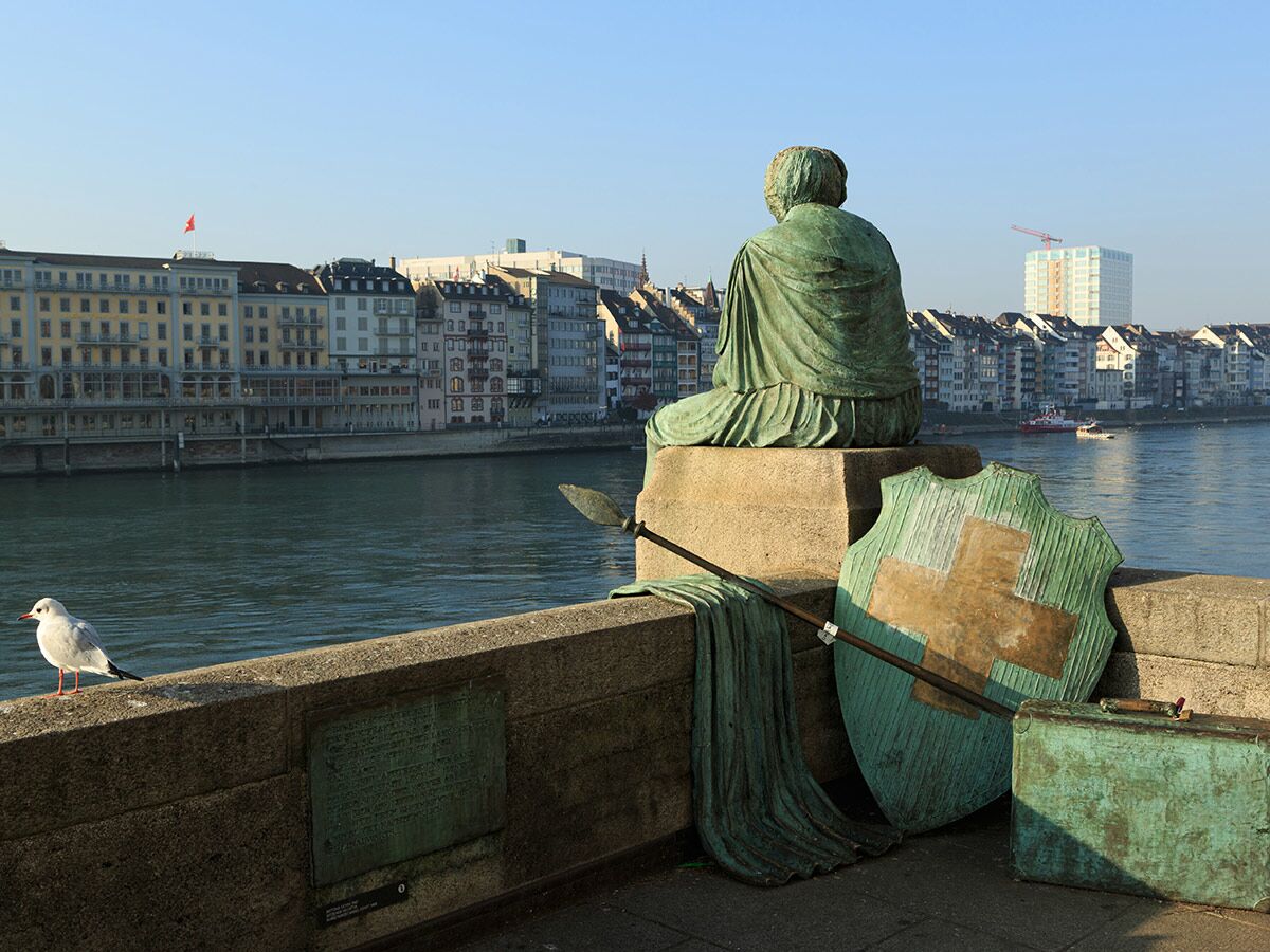 Bronze-Statue mit Schweizer Schild und Möwe am Rheinufer in Basel, Blick auf die historische Stadtkulisse und den Fluss.