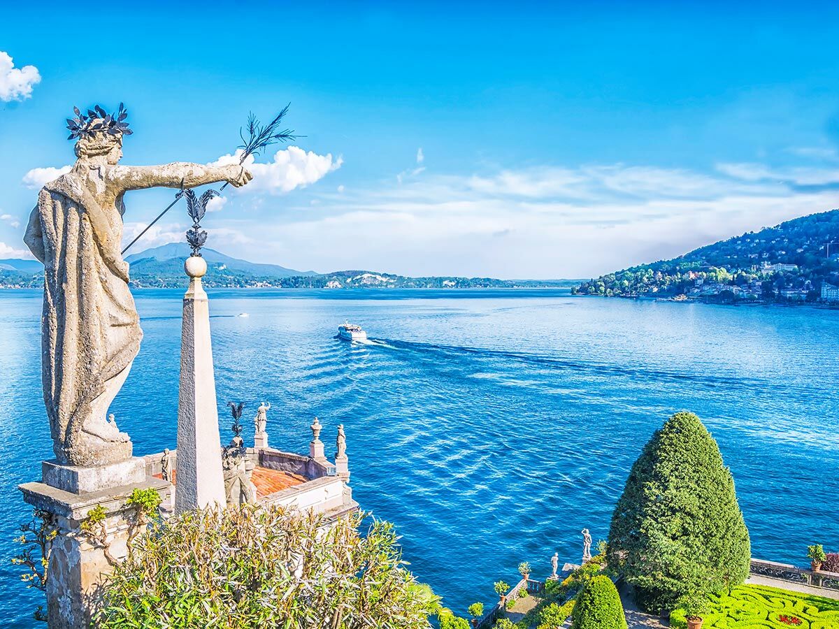 Majestätische Steinstatue mit Lorbeerkranz überblickt den Lago Maggiore, Schiffe und Berge unter blauem Himmel.