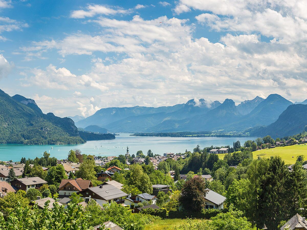 Panoramablick auf den malerischen Wolfgangsee mit Segelbooten, einem Gleitschirm, idyllischem Dorf und Alpenbergen.